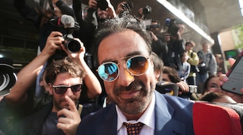 Primer plano de un hombre con barba, traje azul, camisa blanca, corbata y gafas de sol, rodeado por personas con cámaras, micrófonos y un teléfono móvil.