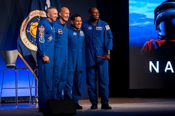 Cuatro hombres con trajes azules de astronauta y parches posan en un escenario. Hay una bandera de NASA y una pantalla con un casco y el texto NASA