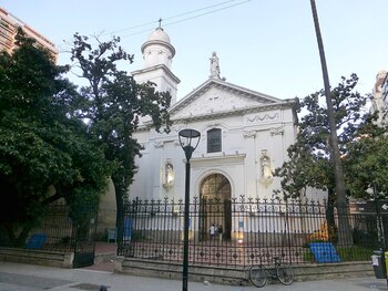 Vista exterior de la Iglesia Santa Catalina y Monasterio, edificio blanco con campanario, estatuas y reja de hierro forjado. Árboles, poste de luz y bicicleta visibles