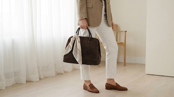 Persona con blazer beige, pantalón blanco y mocasines, sosteniendo un bolso de gamuza oscuro con pañuelo rayado. Detrás hay cortinas blancas y un suelo de madera clara.