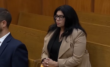 Lubby Navarro, una mujer de cabello oscuro y gafas, sentada en un banco de madera en un tribunal, con un blazer beige y una camisa negra, manos entrelazadas