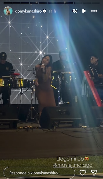 Una mujer canta frente a un micrófono en un escenario iluminado, acompañada por músicos con instrumentos de percusión y teclados