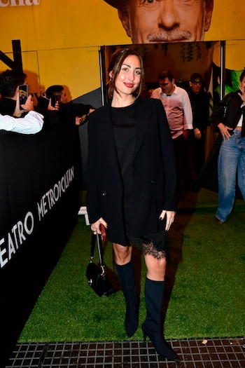 Mujer joven de pie, vestida con blazer negro, vestido con encaje y botas altas, posa en una entrada con césped artificial y una valla que dice 'Teatro Metropolitan'