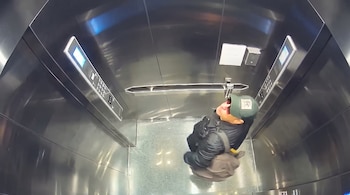 Vista en plano cenital de un hombre con gorra verde, gafas y abrigo dentro de un ascensor con paredes metálicas.
