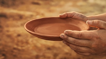 Dos manos sostienen un plato llano de cerámica vacío sobre un fondo desenfocado en tonos ocres y cálidos.