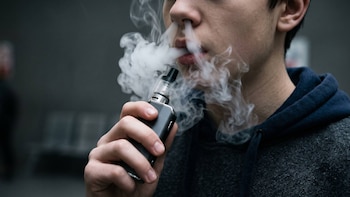 Primer plano de un joven exhalando vapor por boca y nariz. El joven sostiene un cigarrillo electrónico oscuro con la mano derecha