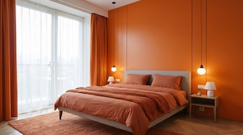 Un dormitorio moderno con paredes naranjas, una cama con ropa de cama naranja y una manta de punto. Hay una ventana grande con cortinas blancas y dos mesitas de noche con lámparas.