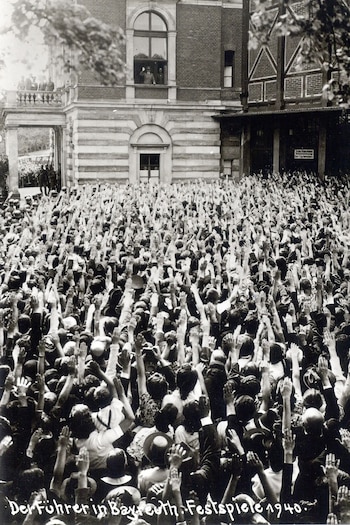 Fotografía en blanco y negro de una gran multitud con los brazos extendidos frente a un edificio de ladrillo; una persona saluda desde una ventana