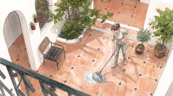 Una mujer trapea un piso de baldosas en un patio, con una mopa. Hay varias plantas en macetas, una silla y una barandilla metálica.