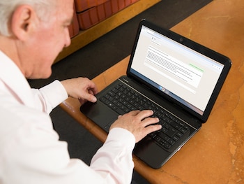 Vista aérea de un hombre mayor con camisa rosa claro usando una Netbook negra sobre una mesa marrón, tecleando con ambas manos