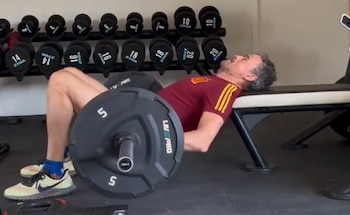 Hombre con camiseta burdeos entrena con barra en un banco de gimnasio, realizando un empuje de cadera. Al fondo, un estante de mancuernas