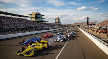 Vista aérea de coches de carreras IndyCar alineados en una pista de carreras asfaltada. Tribunas llenas de público se extienden a los lados, bajo un cielo azul con nubes.