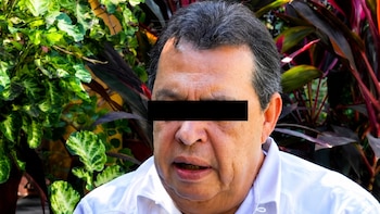 Retrato cercano de un hombre con el área de los ojos censurada. Viste una camisa blanca y tiene cabello oscuro con canas. Fondo con plantas verdes y rojas