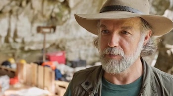 Un hombre con barba canosa, sombrero de ala ancha y chaleco en el interior de una cueva con equipos al fondo