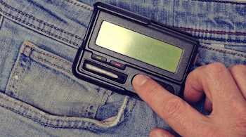 Primer plano de una mano presionando un botón en un beeper negro. El dispositivo se encuentra sobre unos pantalones vaqueros azules.