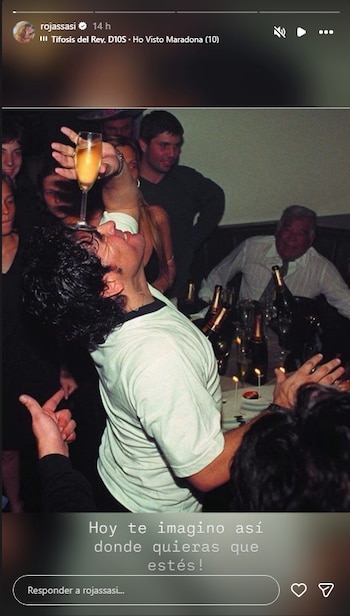 Diego Maradona sostiene una copa sobre su frente en una reunión con varias personas alrededor de botellas y un pastel con velas