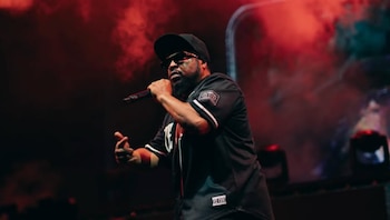Ice Cube viste gorra y camiseta negra mientras sostiene un micrófono en un escenario con humo e iluminación roja de fondo