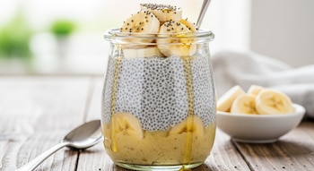 Vaso de cristal con capas de natilla de plátano y semillas oscuras, decorado con rodajas de plátano y miel goteando. Cuchara de metal y tazón con plátano.