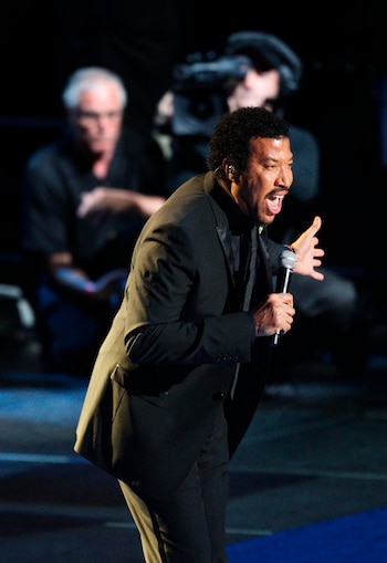 Lionel Richie, vestido con traje oscuro, sostiene un micrófono y gesticula con la mano derecha. El fondo es oscuro con personas desenfocadas