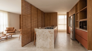 Cocina con isla de mármol, parrilla de cocción, paneles divisores de celosía de madera, refrigerador y una sala contigua.