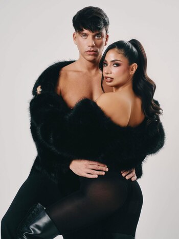 Daniela Celis y Nick Sicaro, él sin camisa con un abrigo de piel negro y ella con medias negras, posan abrazados contra un fondo blanco