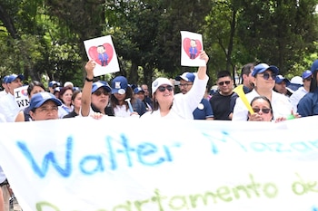 Grupo de personas en una calle con árboles, llevan camisetas blancas, gorras azules, y sostienen carteles de un hombre y una pancarta blanca