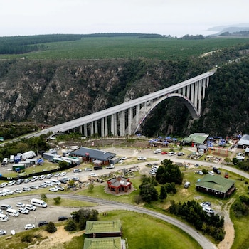 Puente de hormigón en arco sobre un cañón, con vehículos estacionados, pequeñas edificaciones y colinas boscosas alrededor
