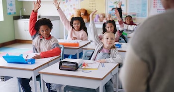 Varios niños en un aula, sentados en escritorios, levantan sus manos hacia un profesor cuya espalda es visible. Hay útiles escolares en las mesas