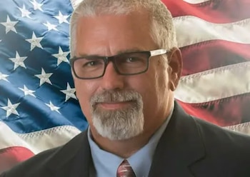 Retrato de un hombre de mediana edad con cabello y barba grises, gafas, traje y corbata, con la bandera estadounidense ligeramente borrosa de fondo