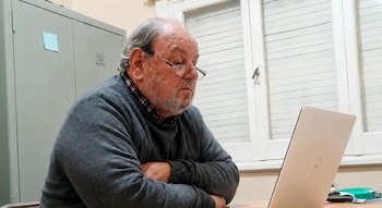 Un hombre con barba y anteojos mira una computadora portátil sobre un escritorio, frente a una ventana blanca y un mueble metálico