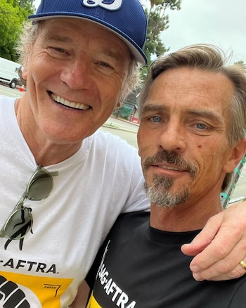 Bryan Cranston con gorra azul y camiseta blanca sonríe junto a Charles Baker, quien viste una camiseta negra