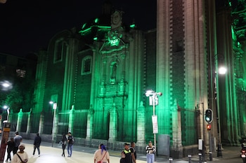 Vista nocturna de la fachada de un edificio colonial en el Centro Histórico de CDMX, iluminado con luz verde vibrante, y personas paseando