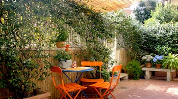 Sillas naranjas y mesa azul en una terraza con suelo de barro, rodeadas de macetas con flores y vegetación bajo un toldo.