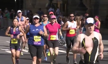 Grupo de personas corriendo en una carrera al aire libre. Corredores usan camisetas, pantalones cortos y dorsales con números. Se observan barreras laterales y público