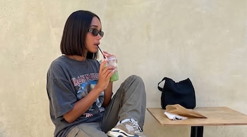 Una mujer con cabello bob, gafas de sol y vestimenta casual, bebe de un vaso. A su lado, una mesa con un bolso negro y una bolsa de papel.