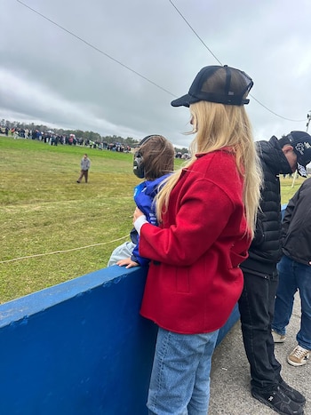 En un circuito, mujeres y niños con auriculares disfrutan del evento. Se ve un coche de carreras azul y una mujer con abrigo rojo en el pit lane
