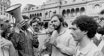 Imagen en blanco y negro de una multitud de hombres frente a un edificio. Luis Brandoni con barba habla por micrófono; Eduardo Blanco a su lado