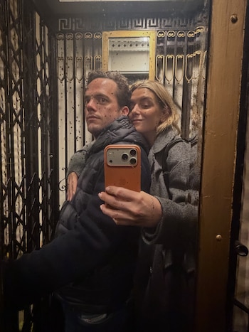 Un hombre y una mujer con manchas oscuras en la piel posan para un selfie en el espejo de un ascensor antiguo con puerta de reja. La mujer sostiene un móvil naranja
