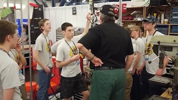 Un hombre adulto de espaldas, con gorra, gesticula ante un grupo de jóvenes que lo observan en un taller o exhibición con herramientas y equipos