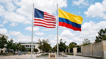 Dos astas de metal sostienen las banderas de Estados Unidos y de Colombia frente a árboles y un edificio bajo un cielo azul con nubes.