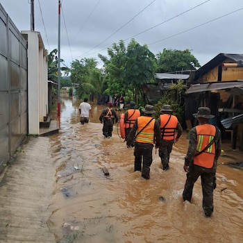 Cinco personas con uniformes militares y chalecos salvavidas naranjas caminan por una calle anegada de agua lodosa. Un hombre de camiseta blanca los precede