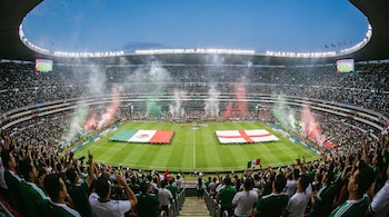 Estadio de fútbol lleno con banderas grandes de México e Inglaterra sobre el césped, público en las gradas, efectos de humo y luz en el campo.