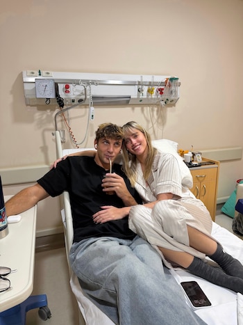 Hombre con camiseta negra y mujer con pijama rayado sentados en cama de clínica. Equipos médicos, mate, teléfono y vasos en la habitación