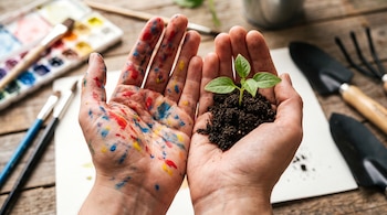 Primer plano de manos: una con pintura de colores, la otra sostiene un brote verde con tierra. Fondo con pinceles, acuarelas y herramientas de jardinería.