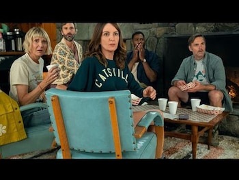 Tina Fey, Tracey Wigfield y Will Forte con expresiones de sorpresa, sentados en una habitación rústica con chimenea de piedra. Hay una mesa de café