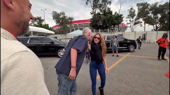 Shakira, con cabello largo y gafas de sol, sonríe junto a un hombre con barba en un estacionamiento. Se observan vehículos y un estadio al fondo