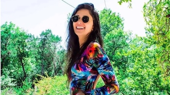 Una mujer con gafas de sol redondas y vestimenta multicolor sonríe frente a una zona arbolada