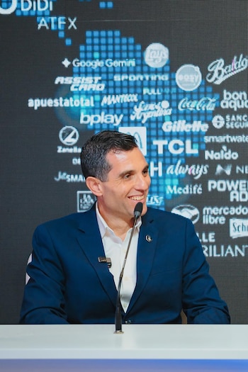 Hombre sonriente con traje azul y camisa blanca habla por un micrófono de pie. Detrás hay una pantalla LED oscura con múltiples logotipos de empresas en azul y blanco