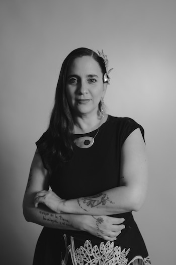 Fotografía en blanco y negro de una mujer de cabello largo, adorno en la cabeza, tatuajes en los brazos y vestido oscuro de brazos cruzados