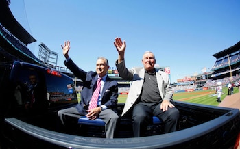 Dos hombres mayores, Bobby Cox y John Schuerholz, saludan desde la parte trasera de una camioneta Chevrolet negra en un estadio de béisbol con un cielo azul claro